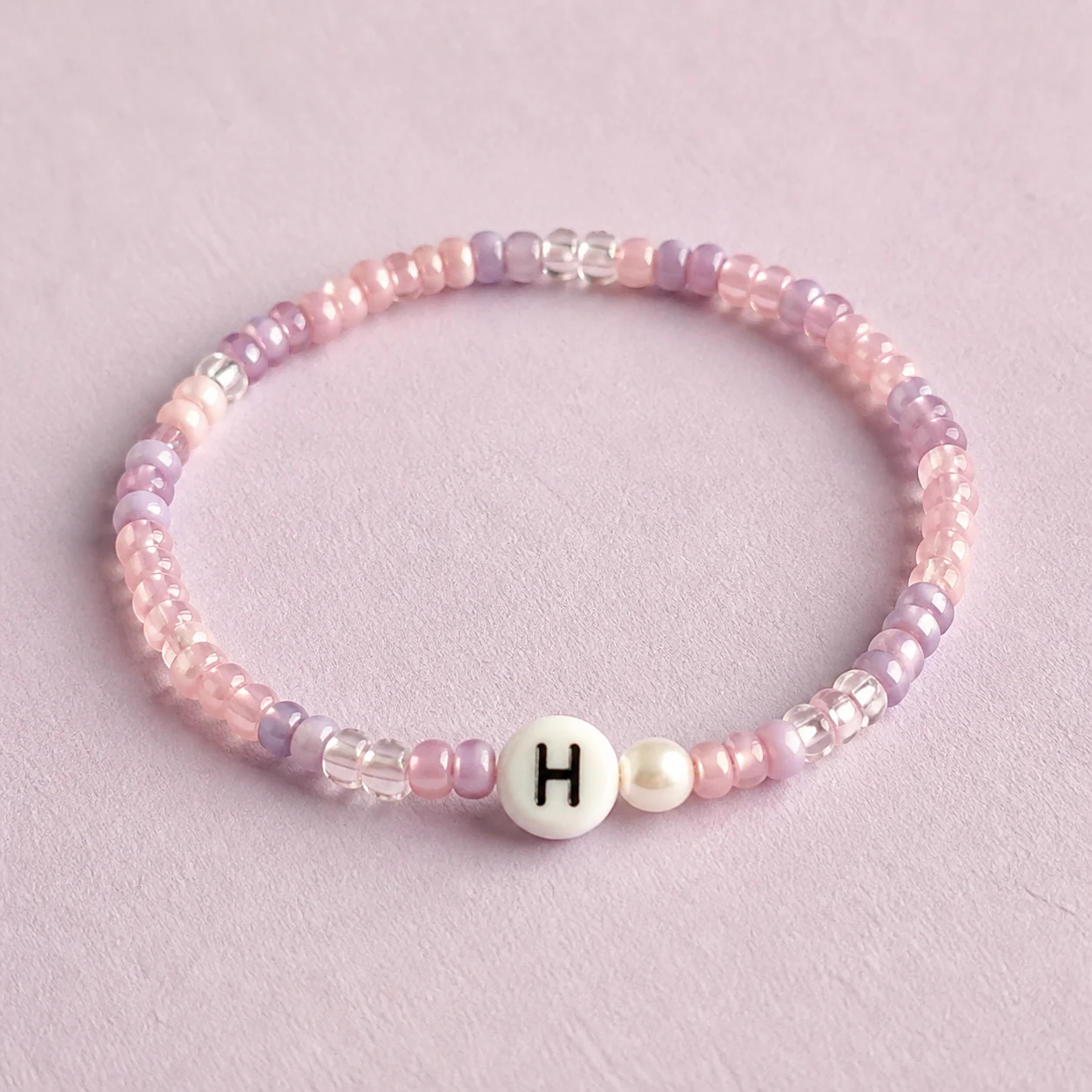 Conceptbeeld van een lichtroze en lila Initial Love-armband met één H en een parelaccent.